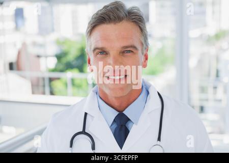 Männlicher Arzt mit weißem Mantel und lächelndem Stethoskop im Klinikkorridor Stockfoto