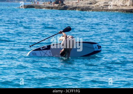 Ein Mann mittleren Alters in seinen 50ern, der versucht, auf einem Stehpaddelbrett auf dem Meer zu balancieren, manchmal sitzend und manchmal ins Wasser fallend. Spaß und Au Stockfoto