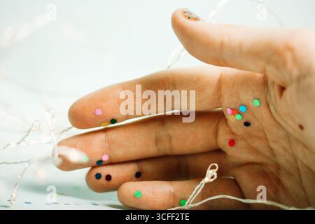 Eine Hand ist mit Glitzer auf der Haut und Lichtern im Hintergrund zu sehen. Eine Hand mit buntem Glitzer, Lichterketten und hellblauem Hintergrund Stockfoto