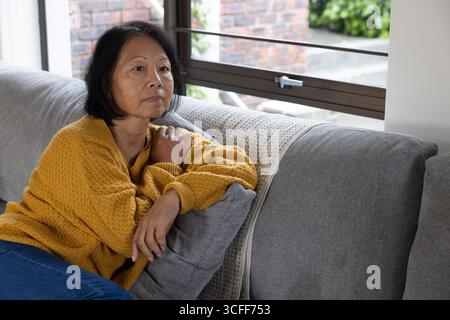 Ältere asiatische Frau, die zu Hause auf der Couch sitzt, am großen Fenster mit strukturiertem Kissen, Kopierraum Stockfoto