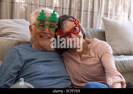 Ein älteres Paar lehnt sich zusammen und lächelt zu Hause auf dem Sofa, trägt eine neue Weihnachtsbrille Stockfoto