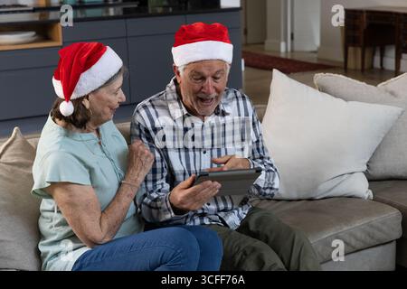 Ein älteres Paar sitzt auf dem Sofa neben Küchenschränken und zeigt auf ein Tablet mit Weihnachtsmützen Stockfoto