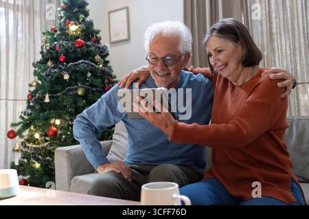 Seniorenpaar mit Pullover, die ein Tablett halten und auf dem Sofa mit Kaffeetasse neben dem Weihnachtsbaum sitzen Stockfoto