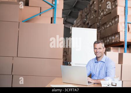 Mann mittleren Alters, der am Schreibtisch im Lagerhaus sitzt, mit Laptop und Whiteboard dahinter, Kopierraum Stockfoto