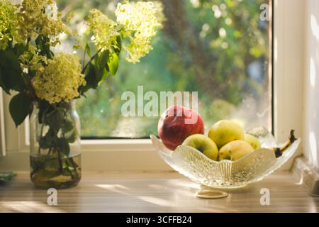 Sonnendurchflutetes Stillleben mit frischem Obst und Blumen auf einer Fensterbank. Ein Stillleben fängt eine sonnenbeschienene Szene mit Früchten in einer Glasschale und Blumen in einer ein ein Stockfoto