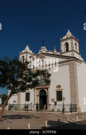 Algarve, Portugal – 2025.06.30: Eine weiße Kirche mit zwei Glockentürmen, verzierten Mauerwerken und einer grünen Eingangstür, die vor einem leuchtend blauen Himmel in A s steht Stockfoto