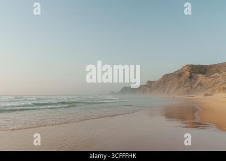 Vila do Bispo, Algarve, Portugal – 2025.07.01 – Goldenes Licht reflektiert auf nassem Sand, während die Wellen unter den Klippen am Praia da Cordoama auf das Ufer treffen. Stockfoto