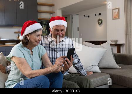 Seniorenpaar, das auf einem grauen Sofa im modernen Wohnzimmer sitzt, trägt rote Santa-Hüte, die das Smartphone berühren Stockfoto