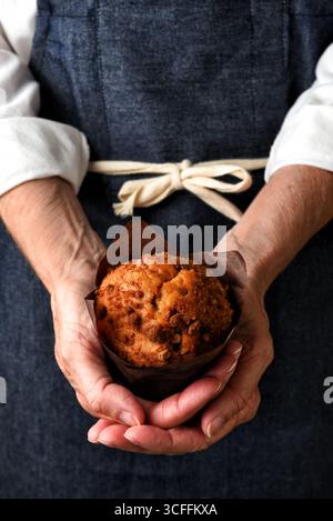 Eine Bäckerin, die einen frisch gebackenen Muffin in der Hand hält. Die Frau trägt eine blaue Denim-Schürze. Stockfoto