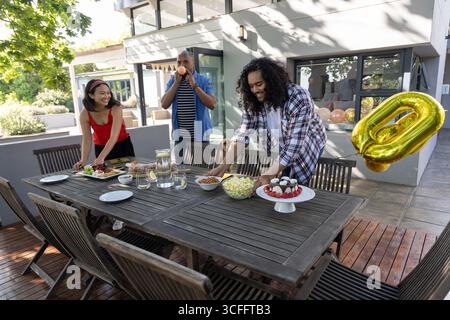 Verschiedene Freunde arrangieren Snacks auf hölzernem Terrassentisch und blasen zu Hause einen orangen Ballon auf Stockfoto