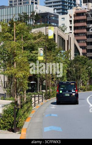TOKIO, JAPAN - 19. August 2025: Straße in den Azabudai Hills mit einem Taxi und einem Banner auf einem Laternenpfosten, der eine Isao Takahata-Ausstellung anpricht. Stockfoto