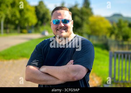 Ein junger Mann mit reflektierender Sonnenbrille und schwarzem T-Shirt steht draußen mit überkreuzten Armen und lächelt selbstbewusst. Stockfoto
