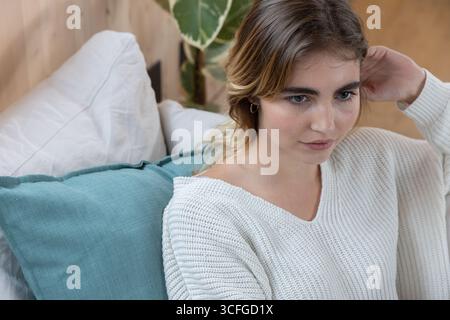 Frau in ihren Zwanzigern, die auf dem Sofa sitzt und einen Strickpullover trägt, der Ohrringe in der Nähe der Zimmerpflanze anpasst Stockfoto