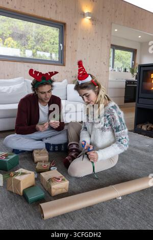 Paare sitzen auf grauem Teppich im Wohnzimmer und wickeln Geschenke mit Schere, Band und Kraftpapier ein Stockfoto