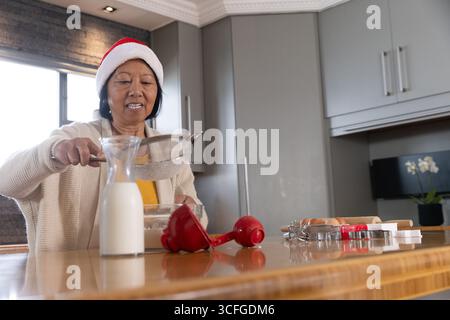 Ältere asiatische Frau mit Weihnachtsmann-Hut, die Mehl durch ein Metallsieb in eine Schüssel an der Küchentheke siebt Stockfoto