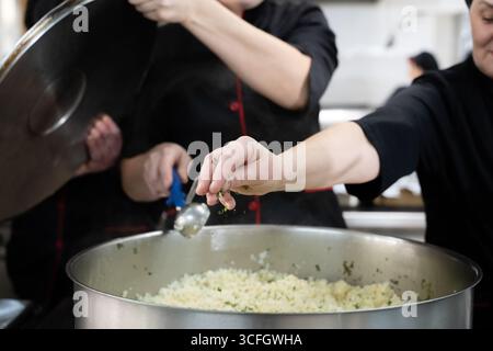 Gruppe von Köchen in schwarzen Uniformen, die Reis in einem großen Topf in einer kommerziellen Küche zubereiten. Weibliche Köche rühren und fügen Kräuter hinzu, während sie als Team arbeiten Stockfoto