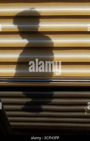 Der scharfe Schatten einer Person wird auf einen gestreiften Rollladen projiziert und bildet eine markante und minimalistische Komposition. Stockfoto