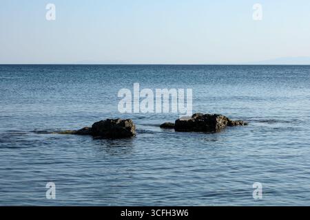 Ruhige Küstenszene mit zwei dunklen Felsen, die aus dem ruhigen Meer auftauchen, mit einem klaren Horizont und einem sanften blauen Himmel im Hintergrund. Stockfoto