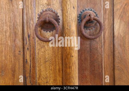 Nahaufnahme einer alten Holztür mit zwei rostigen Eisenringgriffen in symmetrischem, traditionellem Design. Stockfoto
