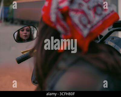 Die junge Bikerin in rotem Bandana trägt Lippenstift auf, während sie ihre Reflexion im Motorradspiegel ansieht. Schönheit und Selbstvertrauen in einem trendigen, urbanen BI Stockfoto