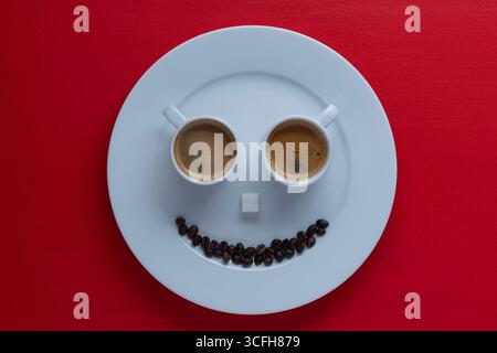 Glückliche Gesichtsform aus zwei Tassen Espresso-Kaffee, Zuckerwürfel und Kaffeebohnen in weißer Platte auf rotem Hintergrund, Nahaufnahme, Draufsicht. Creative com Stockfoto