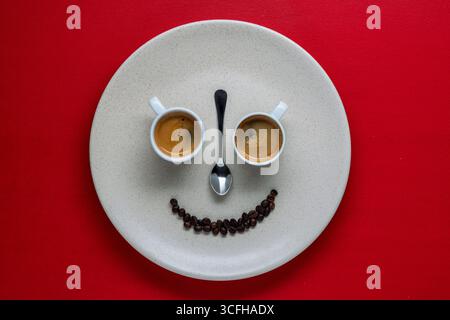 Glückliche Gesichtsform aus zwei weißen Tassen Espresso-Kaffee, Teelöffel und Kaffeebohnen in Teller auf rotem Hintergrund, Nahaufnahme, Draufsicht. Kreative Komposition Stockfoto