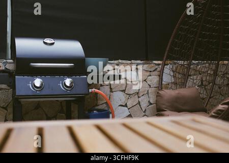 Nahaufnahme eines hölzernen Außentisches mit einem verschwommenen Gasgrill im Hintergrund. Backyard BBQ-Einrichtung bereit für Sommerkochen und gesellige Zusammenkünfte. Stockfoto