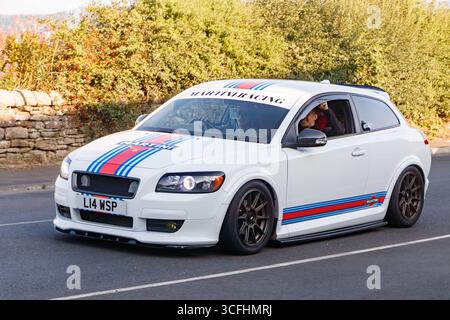 Ein weißer Sportwagen mit Rennstreifen wird auf einer zweispurigen Straße gefahren, umgeben von Grün und einer Steinmauer am Straßenrand. Stockfoto