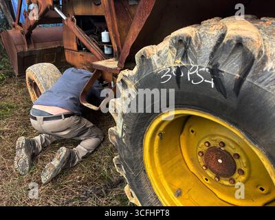 Mann kniet unter dem Fahrgestell des Ladertraktors in schlammiger Umgebung. Kein MR oder PR Stockfoto