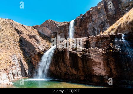 Zweistöckiger Wasserfall in Marcapomacocha, Peru, der über felsige Klippen in einen türkisfarbenen Pool stürzt, umgeben von trockenem Gras und blauem Himmel. Stockfoto