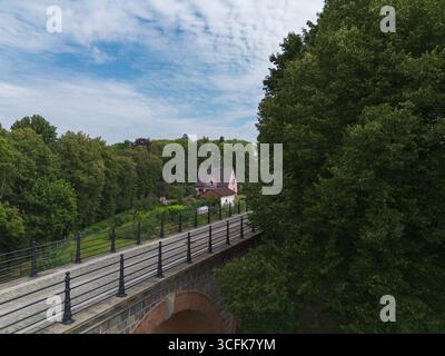 Wunderschöner Blick auf eine alte Eisenbahnbrücke, umgeben von üppigen grünen Bäumen, mit einem bezaubernden rosa Haus im Hintergrund vor einem blauen Himmel Stockfoto