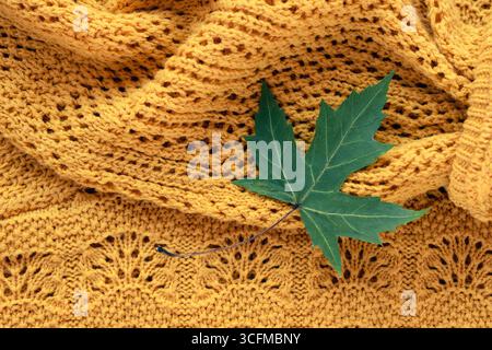 Grünes Ahornblatt, das auf warmem, gelbem Strickmaterial liegt und einen gemütlichen Herbst-Hintergrund schafft. Draufsicht. Stockfoto