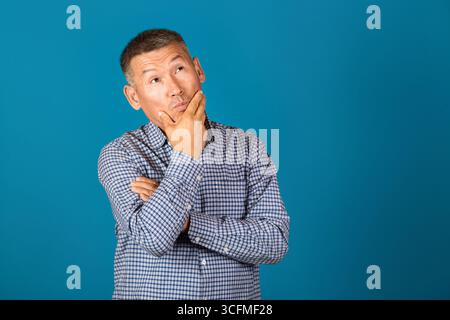 Ein ernster, gutaussehender asiatischer Mann mittleren Alters, der das Kinn vor blauem Hintergrund hält. Stockfoto