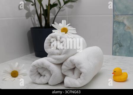 Gerollte weiße Handtücher sind ordentlich auf einer Oberfläche angeordnet, ergänzt durch Blumen und eine verspielte Gummiente. Eine Topfpflanze verleiht einen Hauch von Natur und schafft eine ruhige Atmosphäre. Stockfoto