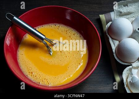 Eier rühren. Rohe Eier in einer roten Schüssel mit einem Schneebesen für Omelette geschlagen, Nahaufnahme. Frühstückszubereitung, rustikaler Stil Stockfoto