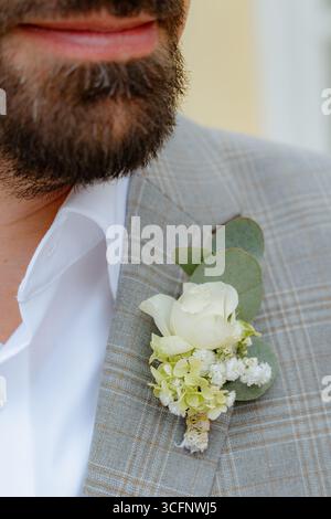 Nahaufnahme einer Bräutigam-Anzugsjacke mit einer ordentlich angeordneten weißen Rosenboutonniere. Das Blumendetail ergänzt das elegante graue Karomuster und verleiht dem Stoff einen besonderen Touch Stockfoto