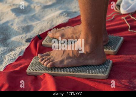 Der Mensch steht auf scharfen Nägeln am tropischen Sandstrand während des Sonnenuntergangs. Männliche Füße auf einem Sadhu-Brett, Nahaufnahme. Konzeptyoga und Meditation Stockfoto