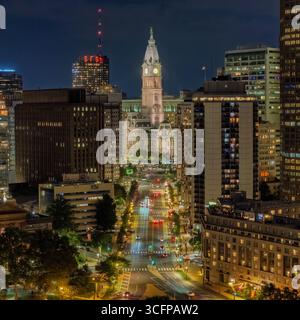 In der lebhaften Stadt Philadelphia, Pennsylvania bei Nacht, befindet sich der große Uhrenturm des Rathauses, das in der Ferne beleuchtet ist und von hohen Bu umgeben ist Stockfoto