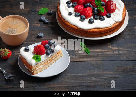 Biskuitkuchen mit Frischkäse und frischen Sommerbeeren auf Holztisch. Stück Kuchen mit Schlagsahne, Erdbeeren und Heidelbeeren und Kaffee. Stockfoto