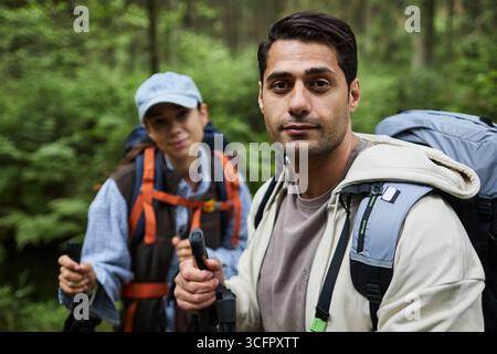Porträt eines jungen Erwachsenen kaukasischen Mannes, der in die Kamera blickt, während er im Wald mit einer jungen Erwachsenen asiatischen Frau, die hinter ihm mit Rucksack und Trekkingstöcken läuft Stockfoto