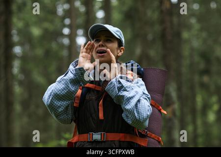Junge Erwachsene kaukasische Frau, die im Wald steht und mit den Händen um den Mund ruft, Rucksack und Mütze trägt und an Outdoor-Aktivitäten inmitten von Bäumen teilnimmt Stockfoto