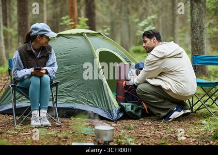 Junge Erwachsene asiatische Frau, die auf Klappstuhl sitzt und mit einem jungen Erwachsenen kaukasischen Mann spricht, der in der Nähe des Zeltes auf dem Waldcamping kniet, beide bereiteten Campingausrüstung vor und sahen sich gegenseitig an Stockfoto