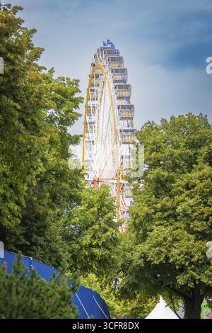 Riesenrad versteckt hinter dichten grünen Bäumen unter blauem Himmel, Olympiapark, München, Deutschland Stockfoto