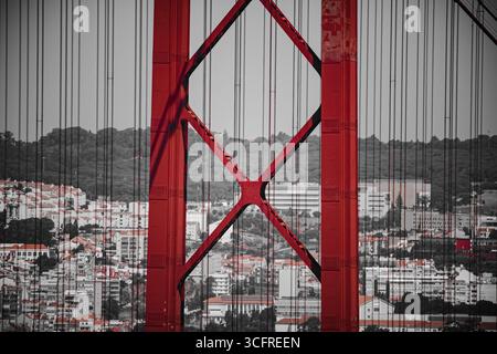 Das Bild zeigt eine Nahansicht einer roten Brückenstruktur, möglicherweise einer Hängebrücke, mit einer Stadt im Hintergrund. Stockfoto