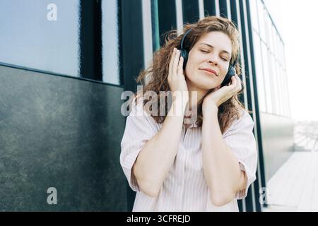 Ein wunderschönes braunhaariges lockiges Mädchen hört draußen Musik auf den Kopfhörern. Stockfoto