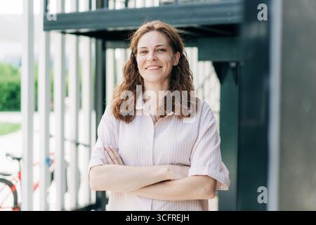 Porträt einer lächelnden europäischen Geschäftsfrau, die die Arme vor dem modernen Gebäude überkreuzt hat. Stockfoto