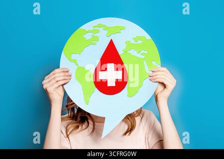 Welthämophilie-Tag. Das Mädchen hält ein Poster mit dem Bild der Erde und einen Tropfen Blut mit einem medizinischen Kreuz. Gesundheitstag, Arzttag Stockfoto