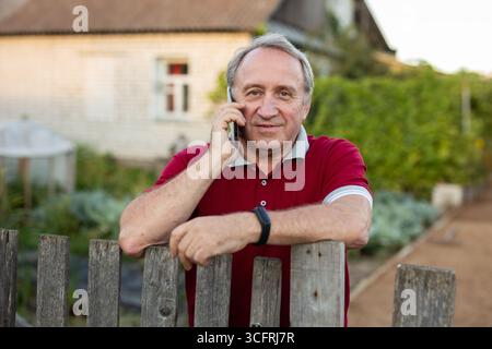 Ein erwachsener Mann, der entspannt neben dem Zaun steht und im Garten telefoniert und lächelt Stockfoto