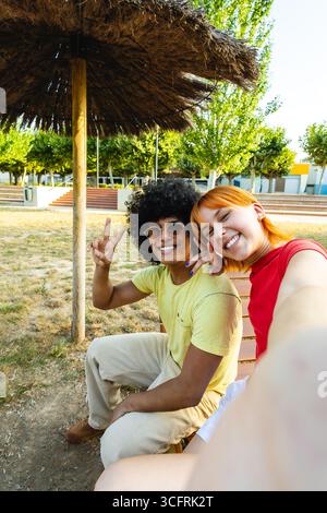 Glückliches Paar, das ein Selfie unter einem Sonnenschirm macht, mit Gesten des Friedenszeichens Stockfoto