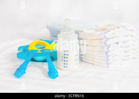 Set aus Babyschale. Milchflasche, Fütterungsknabber, blaues Besteck, Löffel, Gabel, Windeln, Spielzeug, Einwegwindeln auf dem weißen Betthintergrund. Hea Stockfoto